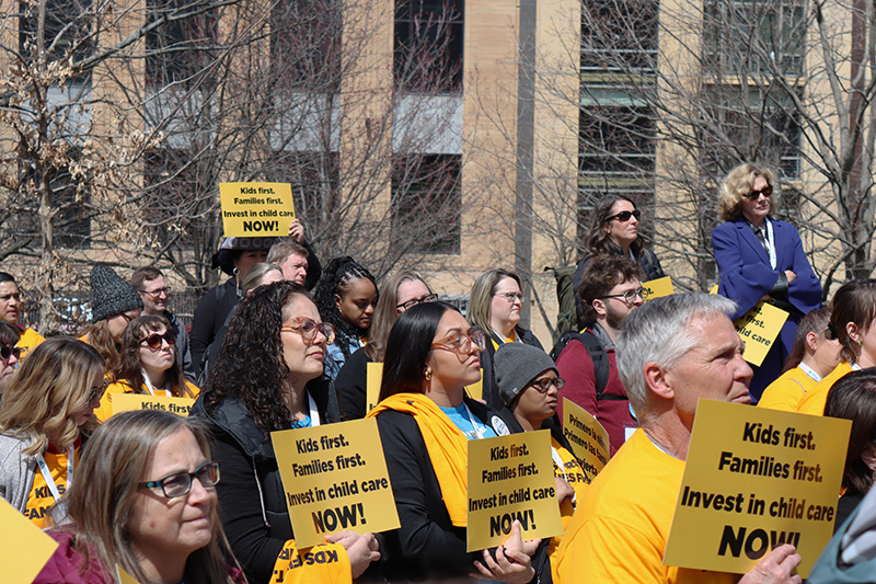 A crowd of people hold yellow signs that read “Kids first. Families first. Invest in child care NOW!” during an outdoor rally.